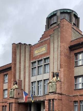 A brick building with an architectural design featuring vertical lines and a central circular glass dome on top. The facade displays two large seated statues flanking the entrance. Below the statues, two flags are visible. The building has multiple large windows and a sign reading 'MUSEUM' in gold letters.