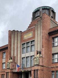 A brick building with an architectural design featuring vertical lines and a central circular glass dome on top. The facade displays two large seated statues flanking the entrance. Below the statues, two flags are visible. The building has multiple large windows and a sign reading 'MUSEUM' in gold letters.