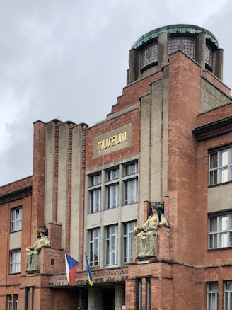 A brick building with an architectural design featuring vertical lines and a central circular glass dome on top. The facade displays two large seated statues flanking the entrance. Below the statues, two flags are visible. The building has multiple large windows and a sign reading 'MUSEUM' in gold letters.