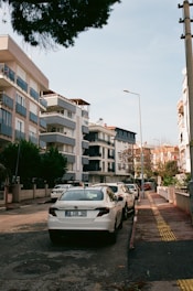 A city street lined with modern apartment buildings and a row of parked cars. The street is clean and has adequate lighting from street lamps. There are small, well-maintained gardens in front of the buildings, and a tree branch is visible at the top of the frame. The sky is clear with a light blue hue.