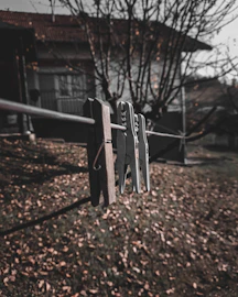 Wooden and metal clothespins are clipped onto a clothesline, with a blurred background of a house and leafless trees. The ground is covered with fallen leaves, suggesting an autumn setting. The focus is sharp on the clothespins, with the rest of the scene gradually fading out of focus.