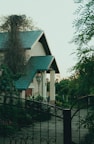 A charming house with a teal roof is partially obscured by lush greenery. The surroundings include tall wrought iron gates and vintage-style street lamps. The garden is flourishing, with various plants and flowers, contributing to a serene outdoor atmosphere.