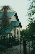 A charming house with a teal roof is partially obscured by lush greenery. The surroundings include tall wrought iron gates and vintage-style street lamps. The garden is flourishing, with various plants and flowers, contributing to a serene outdoor atmosphere.