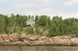A house with a large windowed facade is nestled among dense green trees on a rocky hillside, overlooking a body of calm water. The setting suggests a serene and secluded retreat amidst nature.