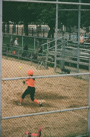 A softball or baseball player wearing an orange uniform and helmet is swinging a bat to hit an incoming ball. The scene is set in a fenced sports field with dirt ground. In the background, there are metal bleachers with a few spectators and equipment such as bats and bags. Tall green trees can be seen beyond the fence.