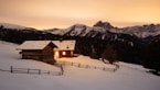 A cozy wooden cabin with lit windows sits on a snow-covered slope, surrounded by wooden fences. In the background, there are forested mountains with snow-capped peaks under a warm, glowing sky.