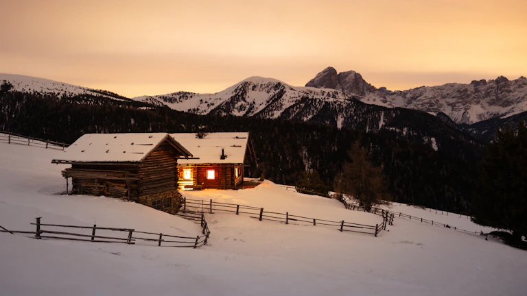 A cozy mountain lodge with warm lights glowing at dusk against the backdrop of snow-capped peaks.