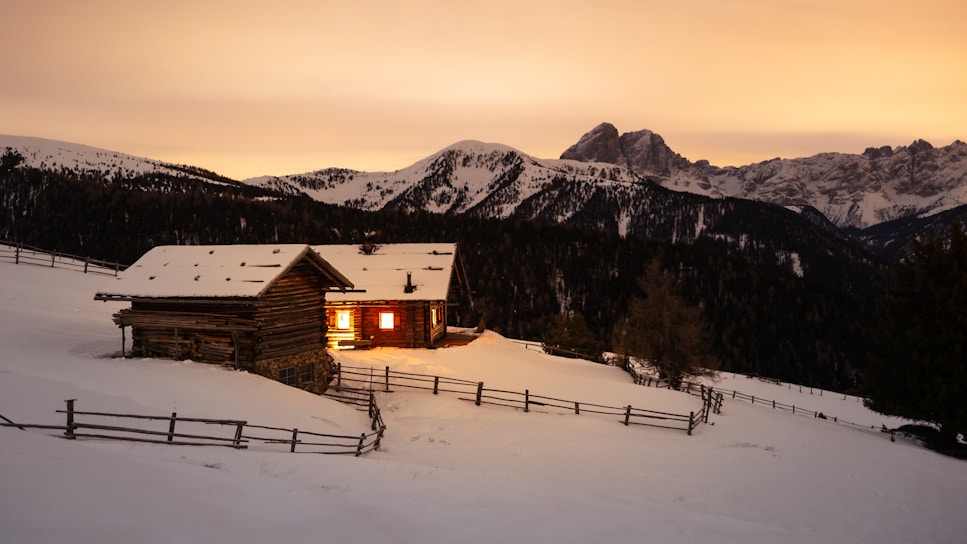 A cozy wooden cabin with lit windows sits on a snow-covered slope, surrounded by wooden fences. In the background, there are forested mountains with snow-capped peaks under a warm, glowing sky.
