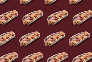 A pattern of hot dogs with black buns, topped with bacon, cheese, and possibly cranberries, arranged on a dark maroon background.