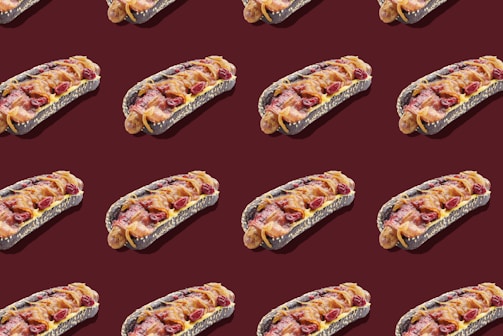 A pattern of hot dogs with black buns, topped with bacon, cheese, and possibly cranberries, arranged on a dark maroon background.
