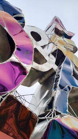 A close-up view of a vibrant, abstract sculpture composed of metallic pieces interspersed with reflective colorful surfaces. Shapes and colors blend together, creating a dynamic and modern aesthetic. The metallic structure is complex, with clean lines and geometric patterns, reflecting the surrounding light and colors.