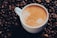 A cup filled with frothy coffee sits among scattered coffee beans, showcasing a rich and inviting beverage. The surface of the coffee displays a light crema.
