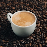 A white coffee cup filled with frothy coffee is set amidst a pile of dark roasted coffee beans.