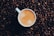 A white coffee cup filled with light brown frothy coffee is centered amidst a backdrop of dark roasted coffee beans.