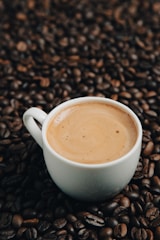 A white cup filled with coffee rests on a surface covered with roasted coffee beans. The coffee has a smooth and creamy appearance, indicating it's freshly brewed.