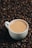 A white cup filled with coffee rests on a surface covered with roasted coffee beans. The coffee has a smooth and creamy appearance, indicating it's freshly brewed.