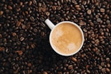 A white cup filled with creamy coffee is surrounded by an abundance of glossy coffee beans. The foam on top of the coffee displays a light swirl pattern.