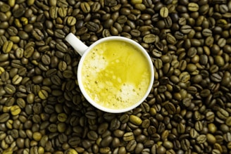 A white cup filled with a yellow frothy beverage is placed on a surface covered with roasted coffee beans.