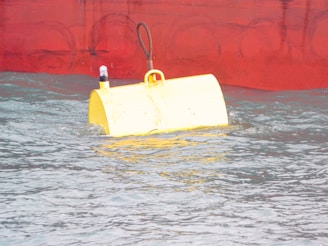 A large, floating yellow buoy is partially submerged in water with a worn red hull of a ship in the background. The buoy has a small light on top and is attached to a cable.