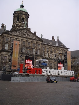 A historical building with a clock tower features prominently, showcasing classic architecture. In front of the building, large colorful letters spell out 'Iamsterdam' with a stage setup and banners for an event. Vehicles are parked in the open square.