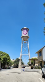 A tall water tower with a large 'WB' logo on its tank stands between low-rise office buildings. The area is lined with trees and parked cars, and the sky is clear and bright blue.
