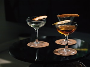 Three cocktail glasses are placed on a round black table, each containing a drink garnished with a twist of citrus peel. The glasses are set on round cork coasters, and the lighting is dim, creating a cozy ambiance.
