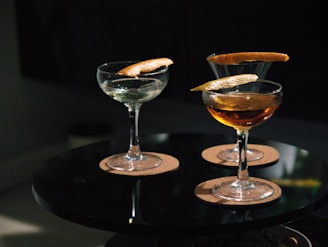 Three cocktail glasses are placed on a round black table, each containing a drink garnished with a twist of citrus peel. The glasses are set on round cork coasters, and the lighting is dim, creating a cozy ambiance.