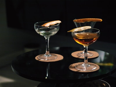 Three cocktail glasses are placed on a round black table, each containing a drink garnished with a twist of citrus peel. The glasses are set on round cork coasters, and the lighting is dim, creating a cozy ambiance.