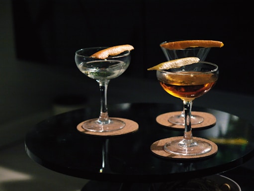 Three cocktail glasses are placed on a round black table, each containing a drink garnished with a twist of citrus peel. The glasses are set on round cork coasters, and the lighting is dim, creating a cozy ambiance.