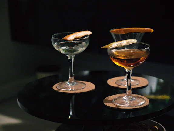 Three cocktail glasses are placed on a round black table, each containing a drink garnished with a twist of citrus peel. The glasses are set on round cork coasters, and the lighting is dim, creating a cozy ambiance.