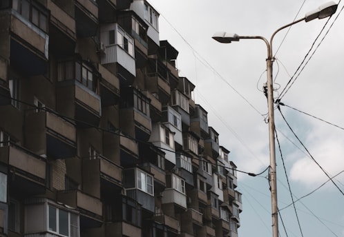 A large, multi-story residential building is shown with numerous balconies and a weathered facade. A streetlight stands in the foreground, with power lines stretching across the image against a cloudy sky.