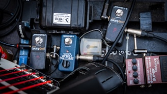 A variety of guitar effects pedals and equipment are organized on a pedalboard. The pedals include a BOSS OC-3 Super Octave and others labeled 'PolyTune' and 'Spectra Comp'. Cables are connected and intertwined, with some loose connectors visible. The background is a textured dark surface, providing contrast to the equipment.