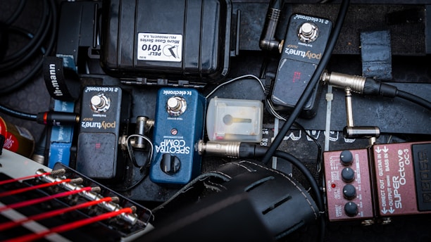 A variety of guitar effects pedals and equipment are organized on a pedalboard. The pedals include a BOSS OC-3 Super Octave and others labeled 'PolyTune' and 'Spectra Comp'. Cables are connected and intertwined, with some loose connectors visible. The background is a textured dark surface, providing contrast to the equipment.