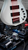 A white bass guitar with bright red strings is positioned above a collection of guitar effect pedals on a pedalboard. The setup includes multiple pedals of varying colors and types, each connected by cables. The background shows a textured black surface.