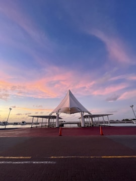 A waterfront scene featuring a modern canopy structure with a pointed roof and surrounding open shelters. The scene is set during sunset, with a beautiful gradient of pink and blue hues in the sky. Two orange traffic cones are placed in front of the structure on a paved surface. In the background, there are boats docked along the water.
