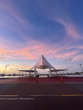 A waterfront scene featuring a modern canopy structure with a pointed roof and surrounding open shelters. The scene is set during sunset, with a beautiful gradient of pink and blue hues in the sky. Two orange traffic cones are placed in front of the structure on a paved surface. In the background, there are boats docked along the water.