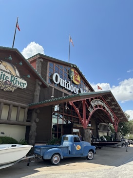 A large outdoor-themed store with a rustic wooden facade, complete with a large arched entrance and flags on top. A vintage blue truck is parked in front, and a white boat is situated nearby. The sky is clear with a few clouds, creating a bright and welcoming atmosphere.