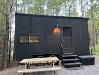 A black, minimalistic tiny house on wheels is situated in a forested area. The house features a small window and a door with a light above it. There's a picnic table in front on a gravel ground. The surrounding is dense with tall trees, and the ambiance feels serene and secluded.