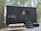 A black, minimalistic tiny house on wheels is situated in a forested area. The house features a small window and a door with a light above it. There's a picnic table in front on a gravel ground. The surrounding is dense with tall trees, and the ambiance feels serene and secluded.