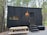A black, minimalistic tiny house on wheels is situated in a forested area. The house features a small window and a door with a light above it. There's a picnic table in front on a gravel ground. The surrounding is dense with tall trees, and the ambiance feels serene and secluded.