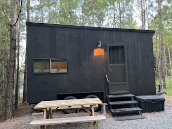 A black, minimalistic tiny house on wheels is situated in a forested area. The house features a small window and a door with a light above it. There's a picnic table in front on a gravel ground. The surrounding is dense with tall trees, and the ambiance feels serene and secluded.