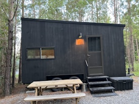 A black, minimalistic tiny house on wheels is situated in a forested area. The house features a small window and a door with a light above it. There's a picnic table in front on a gravel ground. The surrounding is dense with tall trees, and the ambiance feels serene and secluded.