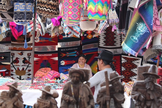 A vibrant market stall featuring a variety of colorful woven textiles, including rugs and bags, displayed alongside handcrafted wooden statues. Two people are sitting and smiling amidst the lively and intricate patterns of the displayed products.