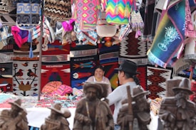 A vibrant market stall featuring a variety of colorful woven textiles, including rugs and bags, displayed alongside handcrafted wooden statues. Two people are sitting and smiling amidst the lively and intricate patterns of the displayed products.