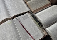 A collection of open books displaying pages with passages and chapters. The text is densely spaced, with some sections highlighted or annotated. A red ribbon bookmark lies across one of the books, and there is a slight shadow cast from an overhead light.