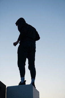 A silhouetted person wearing a hoodie stands atop a concrete block against a clear blue sky. The person appears to be in an upright stance, suggesting a sense of confidence or reflection.