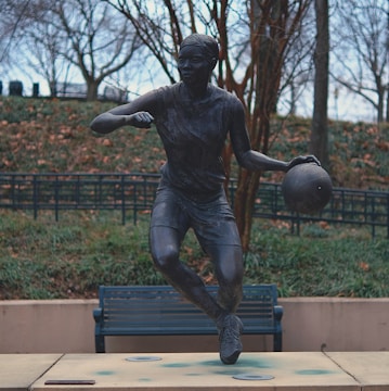 A life-sized bronze statue depicts a person in mid-motion holding a basketball, set in an outdoor park environment surrounded by leafless trees and a park bench in the background.