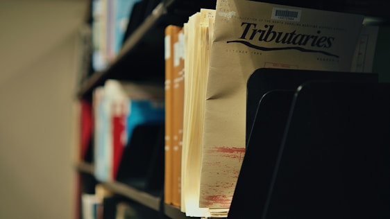 A close-up view of a collection of files or magazines in a library or archive setting. The focus is on a publication titled 'Tributaries' with visible shelves holding other documents in the background. The lighting creates a slightly nostalgic, quiet atmosphere.