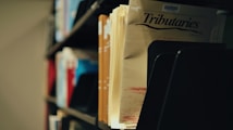 A close-up view of a collection of files or magazines in a library or archive setting. The focus is on a publication titled 'Tributaries' with visible shelves holding other documents in the background. The lighting creates a slightly nostalgic, quiet atmosphere.