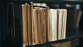 A collection of old, worn folders and files are neatly arranged on a dark shelf. The edges of the papers are yellowed, indicating age, and the surface of the shelf is a deep black, providing contrast to the light-colored documents.